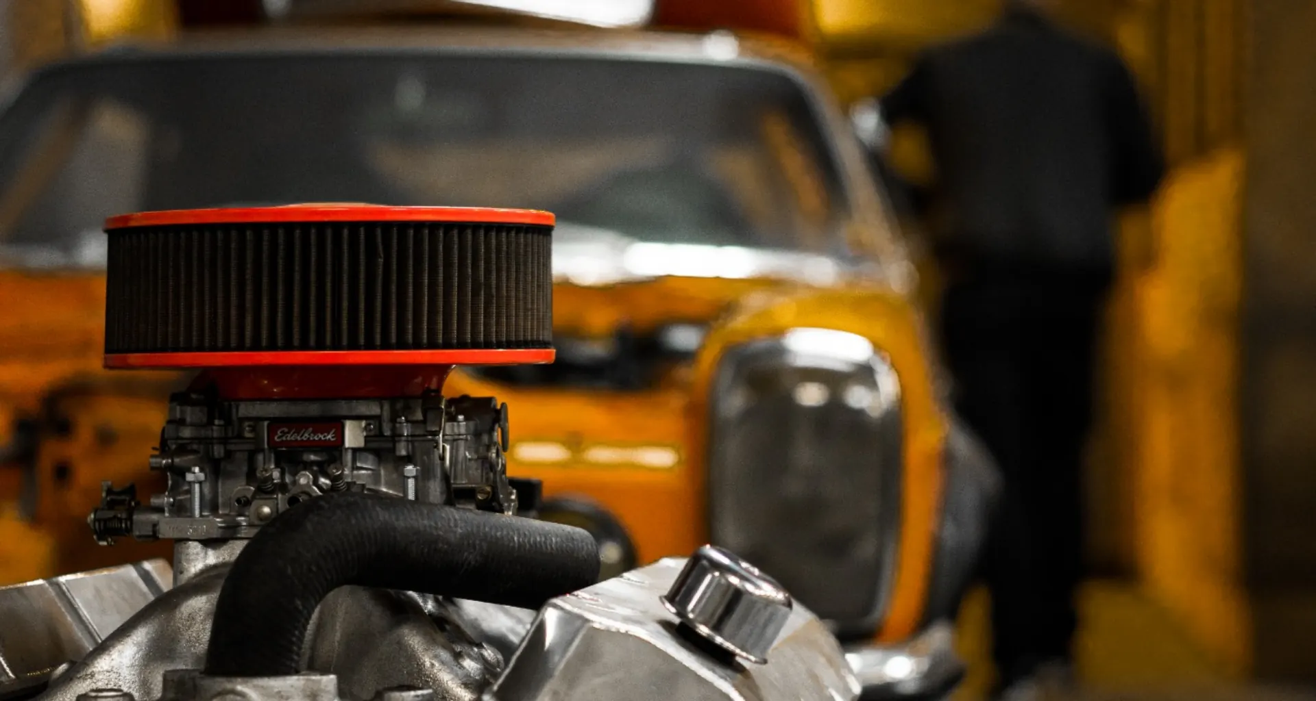 V8-Motor vor einem orangen Oldtimer, im Hintergrund ein Mechaniker